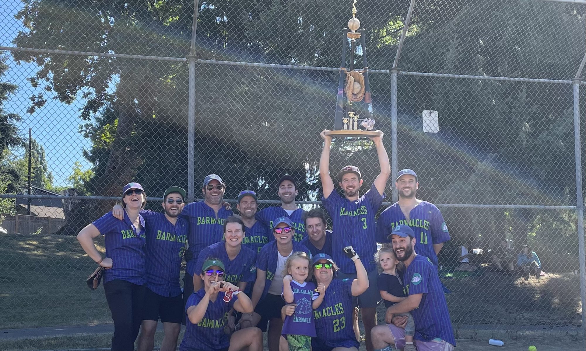 softball players in purples jerseys gather at home plate for a group photo; one player holds a large trophy over their head